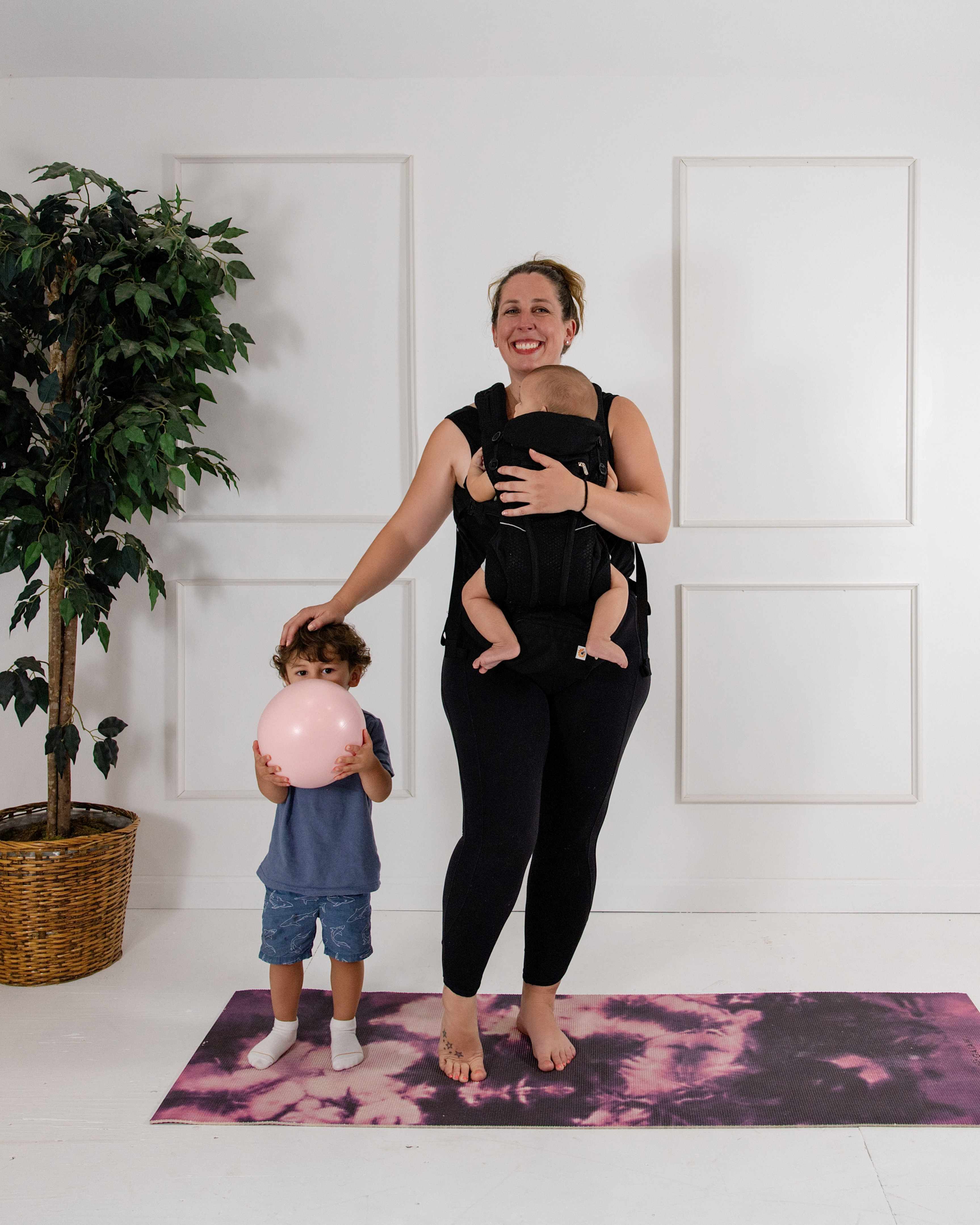Samantha smiling with her baby in a carrier and her toddler holding a pink Pilates ball on a yoga mat