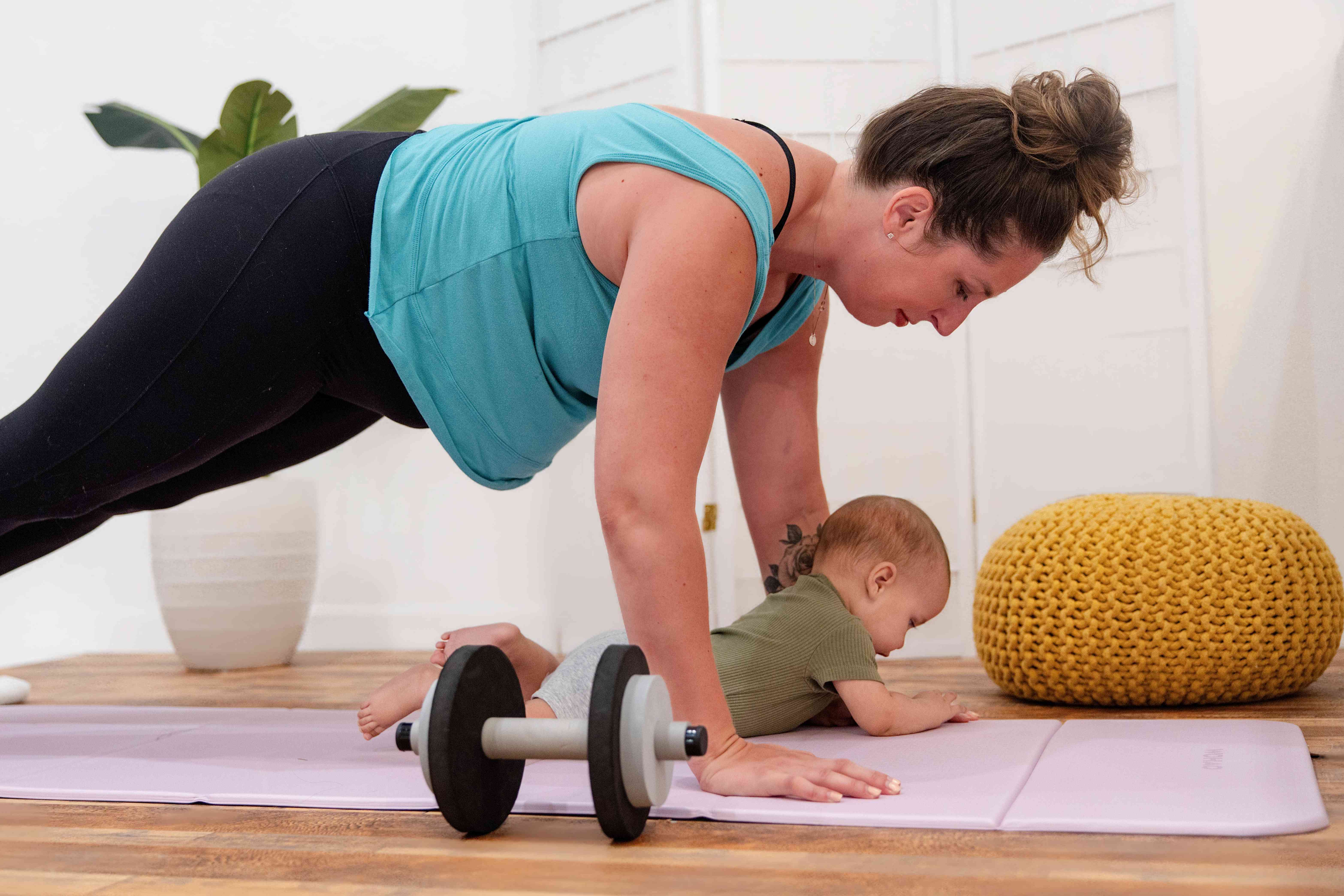 A mom holding a plank on a yoga mat while her baby lies happily beside her